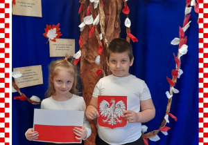 Patrycja i Seweryn w odświętnym ubraniu trzymają w rączkach godło i flagę Polski, w tle dekoracja okolicznościowa